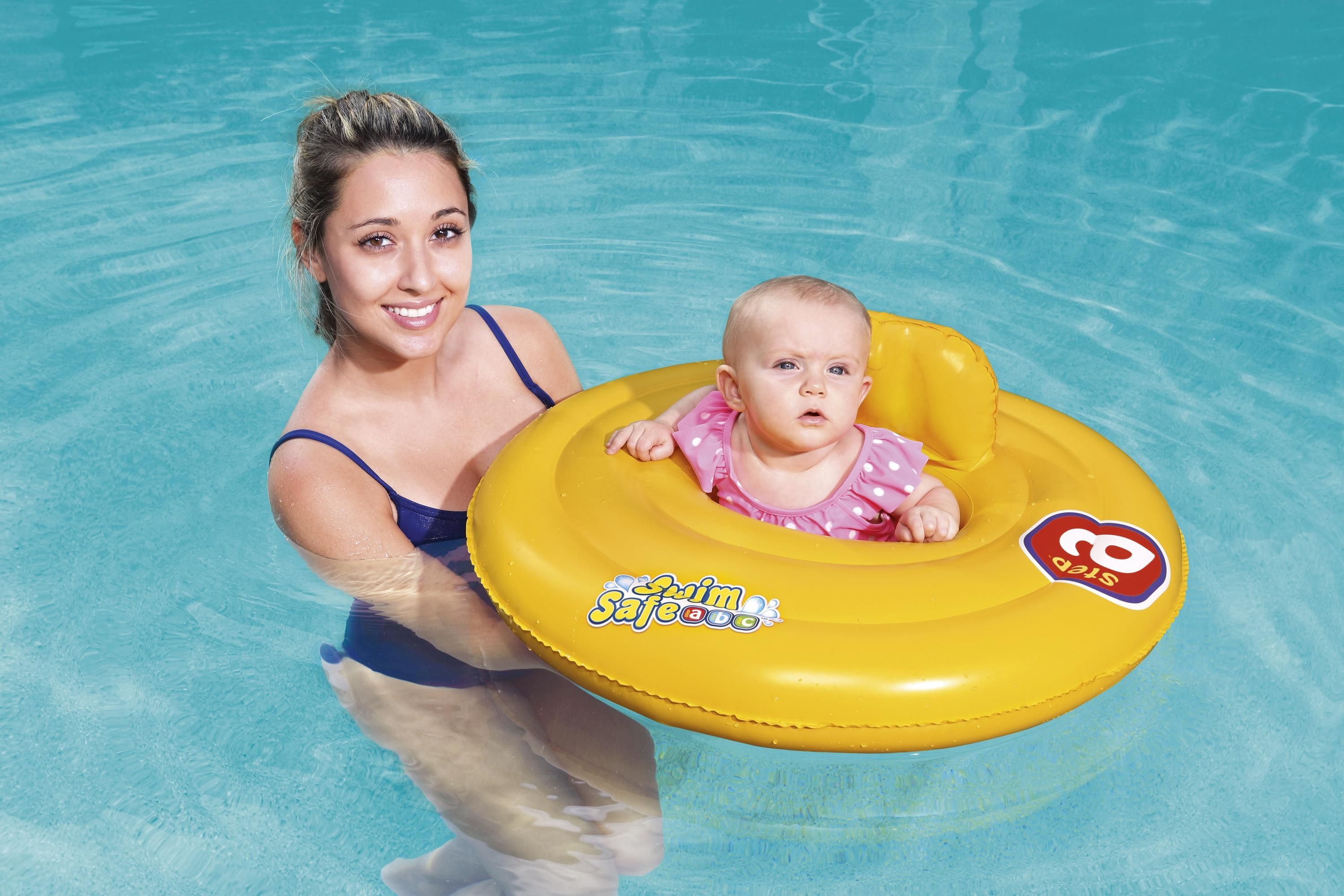 Gul oppblåsbar babyflåte med en baby i rosa prikkete badedrakt, holdt av en kvinne i blå badedrakt.